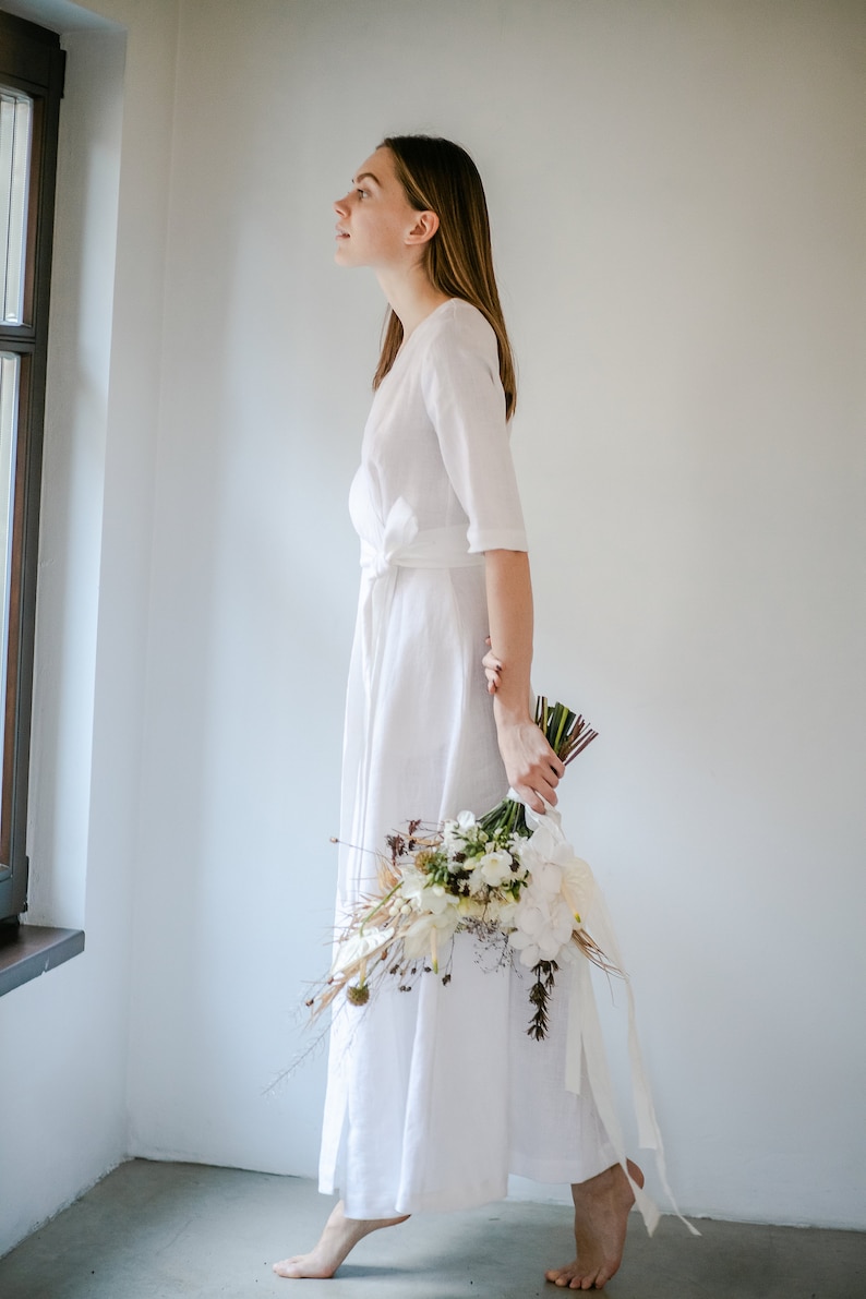 White linen wrap wedding dress with three quarter sleeves, bride holding a bouquet