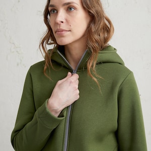 May include: An olive green zip-up hoodie with a hood and a gray zipper. The hoodie is made of a soft, textured fabric. The person in the image is zipping up the hoodie.