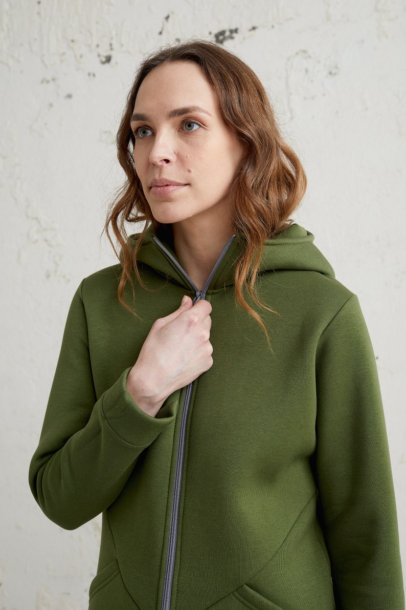 May include: An olive green zip-up hoodie with a hood and a gray zipper. The hoodie is made of a soft, textured fabric. The person in the image is zipping up the hoodie.