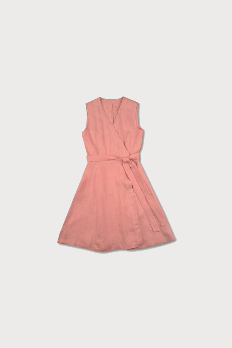 May include: A sleeveless wrap dress in a light pink color. The dress has a V-neckline and a tie belt at the waist.