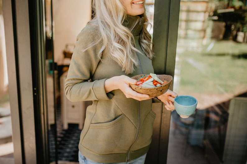 May include: An olive green zip-up jacket with a high collar and front pockets. A wooden bowl filled with sliced vegetables and a small blue ceramic cup are held in the hands. The person is standing near a glass door.