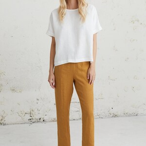 May include: A woman is wearing a white linen top with a relaxed fit and short sleeves. She is also wearing mustard yellow linen pants and brown leather loafers.