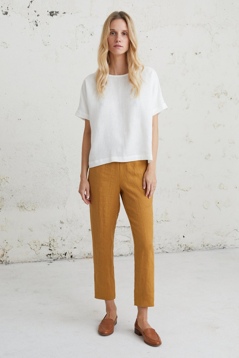 May include: A woman is wearing a white linen top with a relaxed fit and short sleeves. She is also wearing mustard yellow linen pants and brown leather loafers.