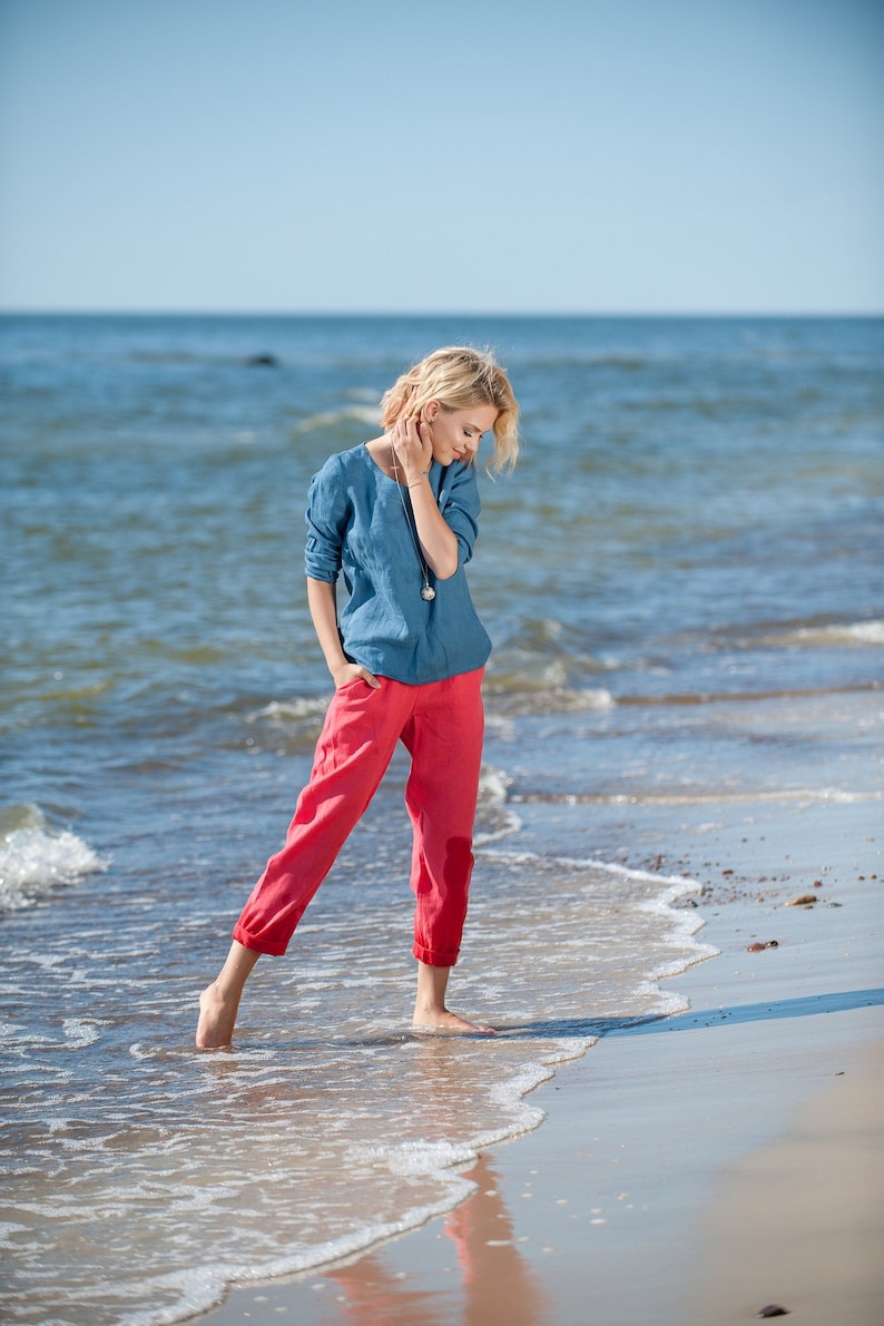 May include: A woman wearing a light blue linen shirt and red linen pants stands on a sandy beach with the ocean behind her.