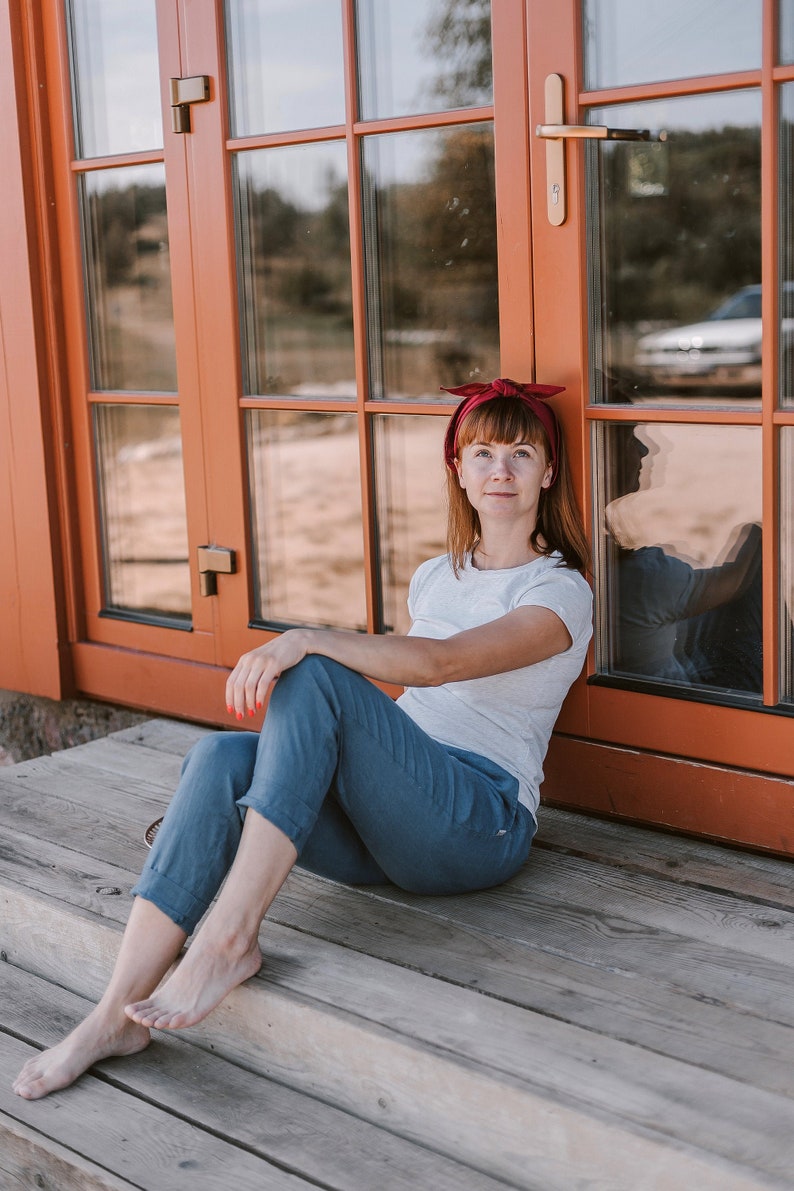 May include: A woman wearing a white t-shirt and blue jeans sits on a wooden porch with a red headband. She is leaning against a wooden door with glass panels.