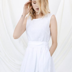 May include: A white linen dress with a sleeveless design and a sash at the waist. The dress has a simple, elegant style.