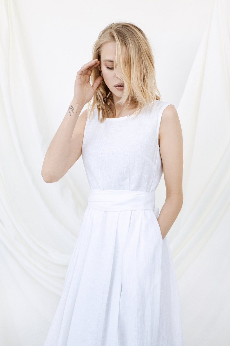 May include: A white linen dress with a sleeveless design and a sash at the waist. The dress has a simple, elegant style.