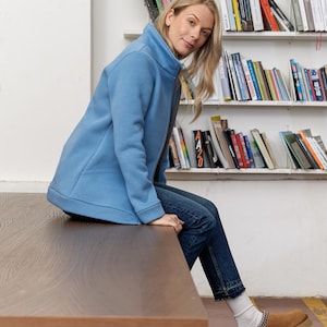 May include: A light blue zip-up jacket with a high collar, paired with blue jeans and brown suede shoes. The jacket has a simple, clean design. The shoes have a chain detail around the ankle. The person is sitting on a wooden table.
