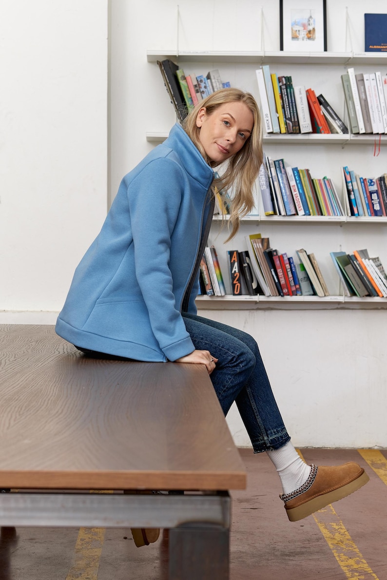 May include: A light blue zip-up jacket with a high collar, paired with blue jeans and brown suede shoes. The jacket has a simple, clean design. The shoes have a chain detail around the ankle. The person is sitting on a wooden table.