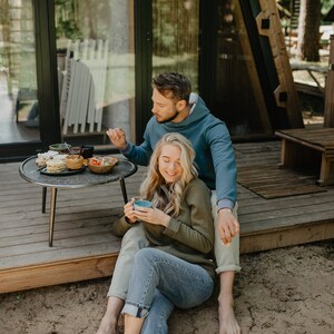 May include: A couple enjoying breakfast outdoors. A small round table holds plates of food and bowls of fruit. The woman is holding a blue mug. They are sitting on a wooden deck in front of a cabin.
