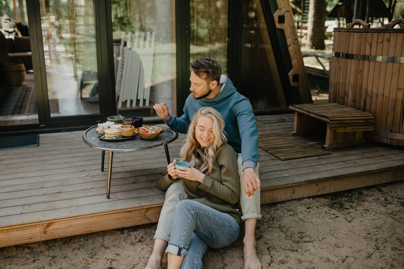 May include: A couple enjoying breakfast outdoors. A small round table holds plates of food and bowls of fruit. The woman is holding a blue mug. They are sitting on a wooden deck in front of a cabin.