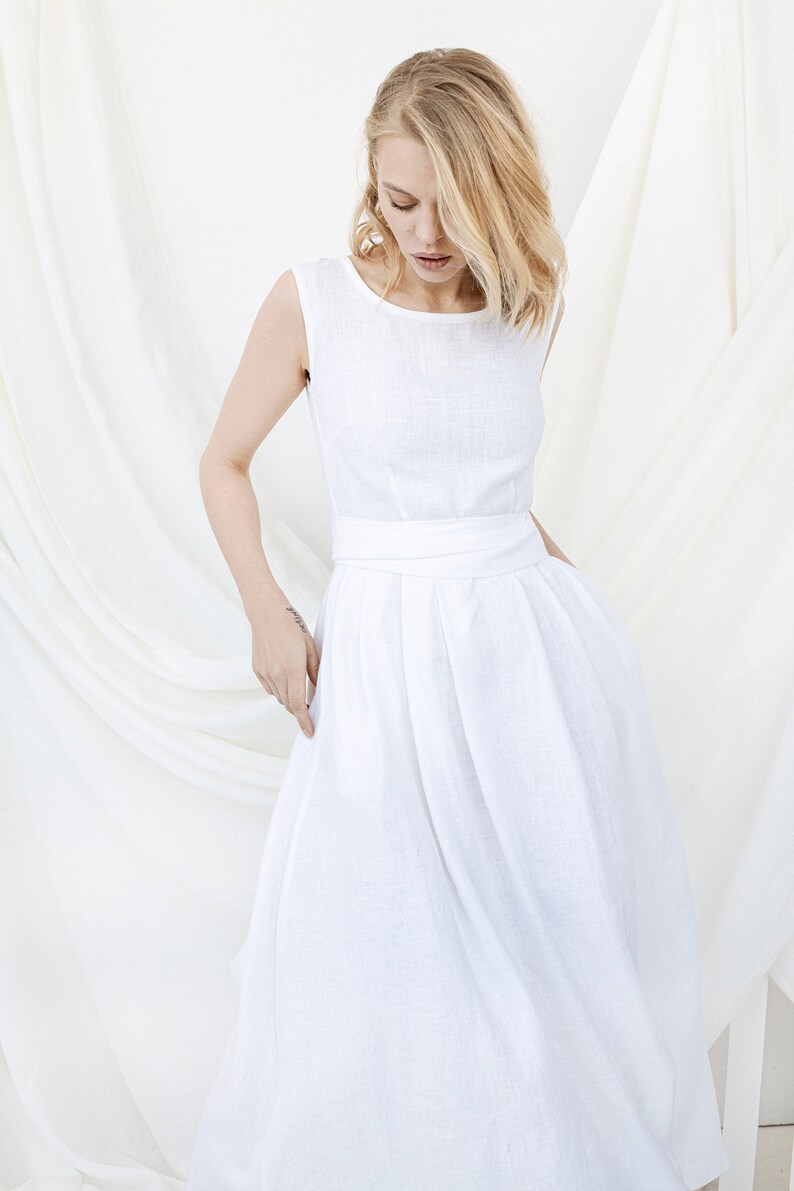 May include: A white linen dress with a sleeveless bodice and a gathered skirt. The dress has a simple, elegant design and is perfect for a summer wedding or other special occasion.