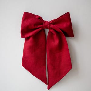 May include: A large, vibrant red bow made of fabric. The bow is centered and has long, flowing tails. The material appears to have a slight texture. The bow is against a plain white background, highlighting its color and shape.
