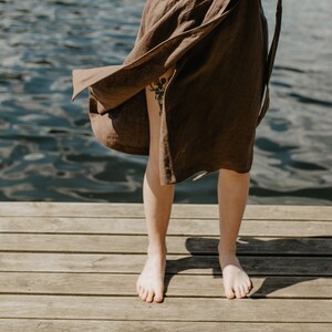 May include: A person standing on a wooden dock wearing a brown linen dress with the skirt blowing in the wind. The person is barefoot and the water is visible in the background.