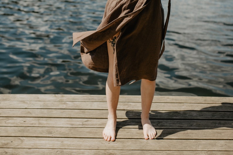 May include: A person standing on a wooden dock wearing a brown linen dress with the skirt blowing in the wind. The person is barefoot and the water is visible in the background.