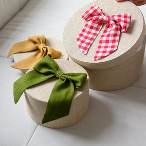 May include: Three round gift boxes with fabric bows. One box has a red and white gingham bow, another has a mustard yellow bow, and the third has a green bow. The boxes are beige and have a textured appearance.