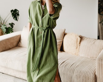 May include: A woman wearing a green linen shirt dress with a belt. She is standing in front of a couch with a light brown fabric.