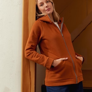 May include: A rust-colored zip-up hoodie with a gray zipper and a hood. The hoodie has two front pockets and is worn with blue jeans. The person is leaning against a wooden door frame.