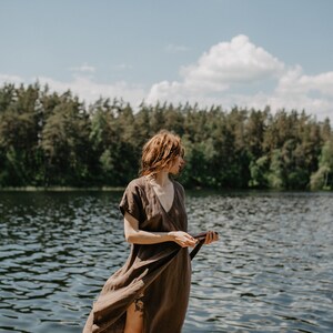 May include: A woman in a brown linen dress stands by a lake with a forest in the background.