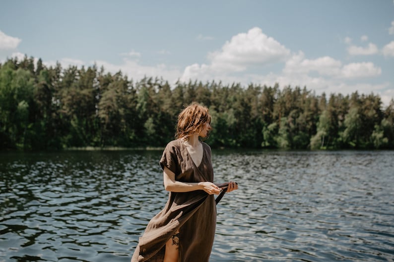 May include: A woman in a brown linen dress stands by a lake with a forest in the background.