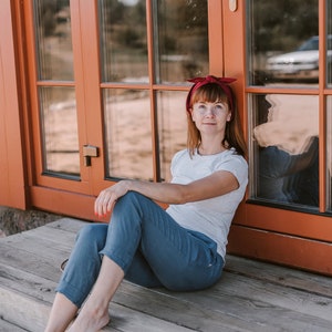 May include: A woman wearing a white t-shirt and blue jeans sits on a wooden porch with a red headband. She is leaning against a wooden door with glass panels.