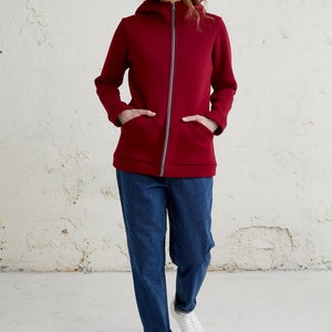 May include: A burgundy zip-up hoodie with a hood and front pockets. The jacket has a gray zipper and is paired with blue jeans and white sneakers. The model is standing in front of a white wall.