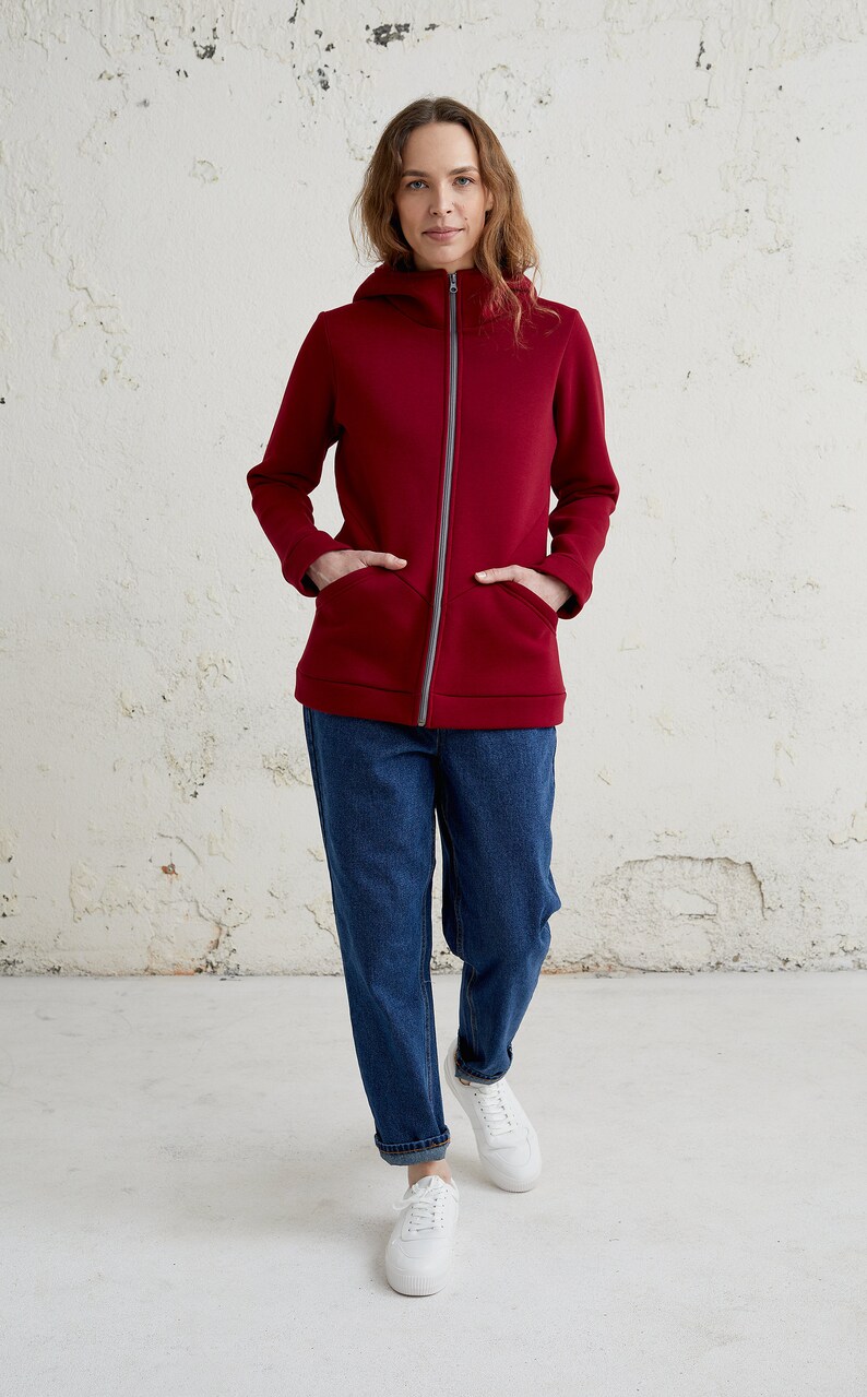 May include: A burgundy zip-up hoodie with a hood and front pockets. The jacket has a gray zipper and is paired with blue jeans and white sneakers. The model is standing in front of a white wall.
