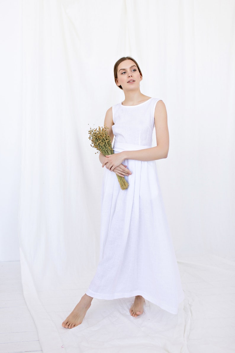 May include: A woman in a white linen dress holds a bouquet of dried flowers. The dress has a simple, sleeveless design and a flowing skirt.
