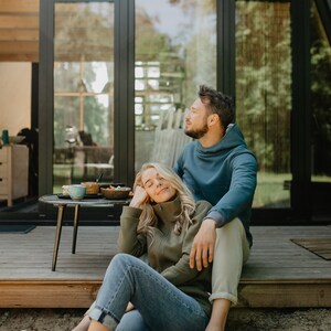 May include: A couple relaxes outdoors, seated on a wooden deck. The woman wears a green zip-up jacket and blue jeans, while the man wears a blue hoodie. A small table holds food and drinks. The scene is set against a backdrop of a modern cabin with large windows.