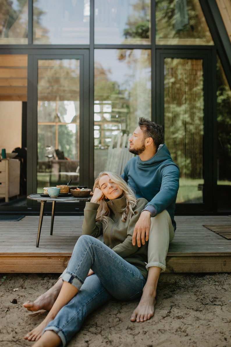 May include: A couple relaxes outdoors, seated on a wooden deck. The woman wears a green zip-up jacket and blue jeans, while the man wears a blue hoodie. A small table holds food and drinks. The scene is set against a backdrop of a modern cabin with large windows.