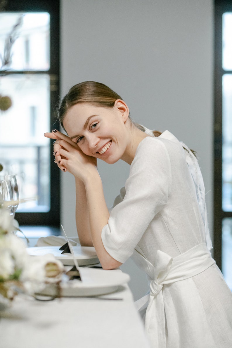 Bride in a white linen wrap dress with a bow at the back, smiling at a table