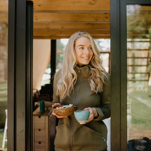 May include: A woman wearing a green zip-up jacket and blue jeans holds a small wooden bowl with salad and a blue ceramic cup. The woman is standing in a doorway with glass panels, looking to the side.
