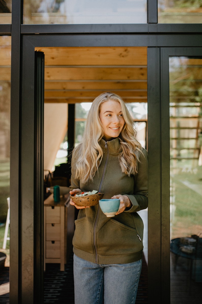 May include: A woman wearing a green zip-up jacket and blue jeans holds a small wooden bowl with salad and a blue ceramic cup. The woman is standing in a doorway with glass panels, looking to the side.