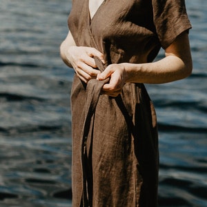 May include: A brown linen wrap dress with short sleeves and a tie belt. The dress is worn by a person standing near a body of water.