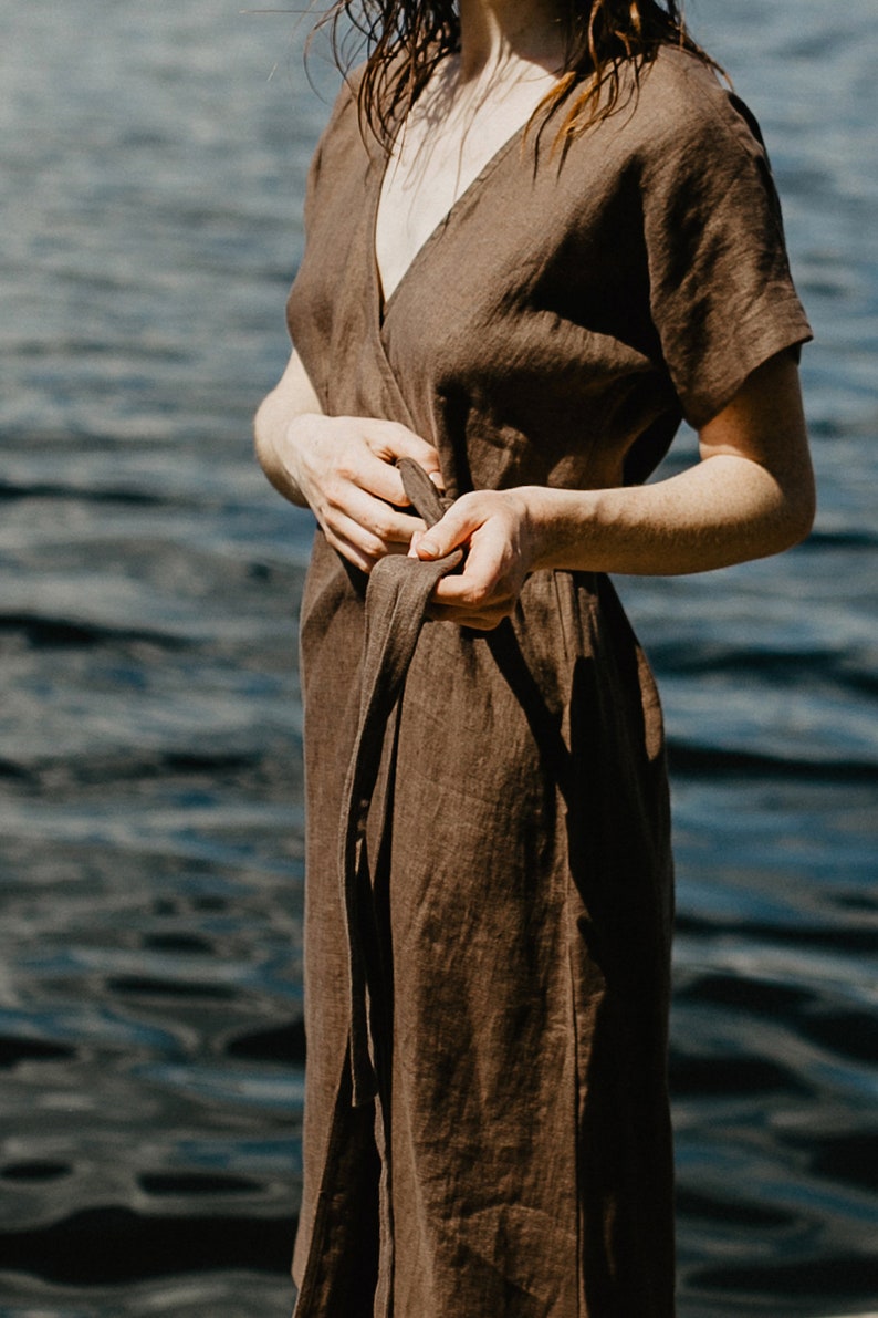 May include: A brown linen wrap dress with short sleeves and a tie belt. The dress is worn by a person standing near a body of water.