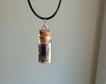 May include: A small glass vial pendant filled with purple and black gemstones, sealed with a cork stopper. The vial hangs from a black cord necklace with a silver-colored metal loop.