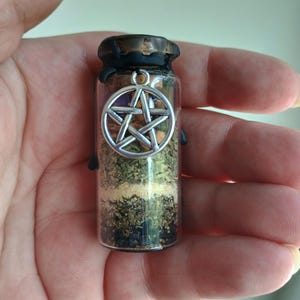 May include: A small glass vial filled with layers of herbs and grains, topped with a dark cap and a silver pentacle charm. The vial is held in a hand, showcasing the layered contents and the decorative charm.
