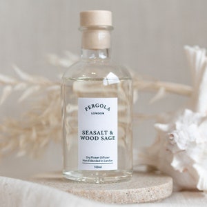Könnte beinhalten: Eine klare Glas-Diffusorflasche mit einem Holzstopfen, beschriftet mit "PERGOLA LONDON SEASALT & WOOD SAGE". Die Flasche enthält eine klare Flüssigkeit und steht auf einem kleinen, runden, beigen Stein. Die Flasche ist 100 ml.