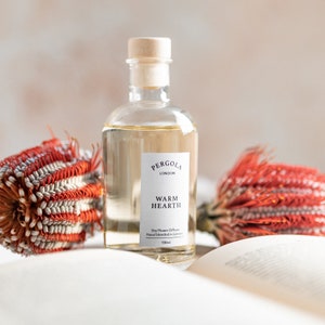 May include: A clear glass bottle of "Warm Hearth" dry flower diffuser from Pergola London, with a wooden stopper. The bottle contains a light amber liquid and is accompanied by red and white dried flowers. The bottle is 100ml.