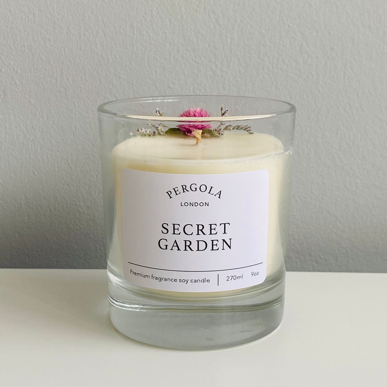 Puede incluir: Un portavelas de cristal transparente con una vela blanca. La vela tiene una etiqueta que dice "SECRET GARDEN" y est&aacute; rematada con flores secas. La vela es de 270 ml y 9 oz.