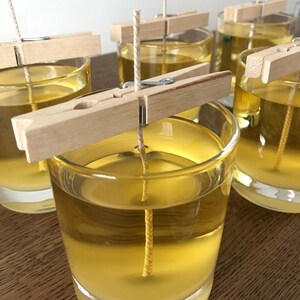 Puede incluir: Primer plano de varios portavelas de cristal transparente llenos de l&iacute;quido amarillo y una mecha. Se utilizan pinzas de madera para sujetar las mechas. Las velas est&aacute;n sobre una superficie de madera.