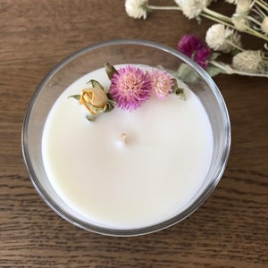 Puede incluir: Un portavelas de cristal transparente lleno de cera blanca. La vela est&aacute; decorada con flores rosas y moradas secas y una peque&ntilde;a rosa amarilla seca. Una mecha de madera est&aacute; centrada en la cera.