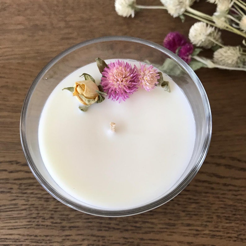 Puede incluir: Un portavelas de cristal transparente lleno de cera blanca. La vela est&aacute; decorada con flores rosas y moradas secas y una peque&ntilde;a rosa amarilla seca. Una mecha de madera est&aacute; centrada en la cera.