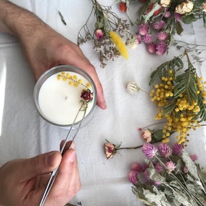 Puede incluir: Un portavelas de cristal transparente con una vela blanca en su interior. Una persona utiliza unas pinzas para colocar flores secas sobre la vela. Flores secas en amarillo, rosa y blanco est&aacute;n esparcidas alrededor de la vela.
