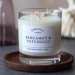 Puede incluir: Un portavelas de cristal transparente con una vela blanca. La vela tiene una etiqueta que dice "BERGAMOT & PATCHOULI" y "PERGOLA LONDON". La vela está sobre una pequeña bandeja redonda de madera. La vela pesa 220g.