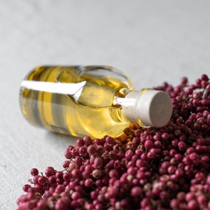 May include: A clear glass bottle filled with yellow liquid, lying on its side. The bottle has a white stopper. A cluster of small, round, red berries surrounds the bottle, creating a natural, rustic aesthetic.