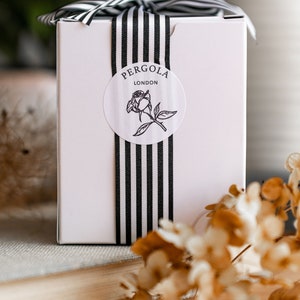 Puede incluir: Una caja de regalo rosa claro con una cinta y lazo a rayas blancas y negras. Una pegatina redonda con el texto "PERGOLA LONDON" y una ilustraci&oacute;n de rosa est&aacute; centrada en la caja. La caja est&aacute; sobre un libro y una superficie de madera.