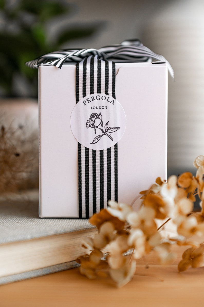 Puede incluir: Una caja de regalo rosa claro con una cinta y lazo a rayas blancas y negras. Una pegatina redonda con el texto "PERGOLA LONDON" y una ilustraci&oacute;n de rosa est&aacute; centrada en la caja. La caja est&aacute; sobre un libro y una superficie de madera.