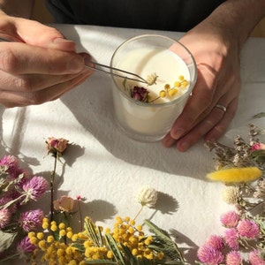 Puede incluir: Una vela de vidrio transparente llena de cera blanca, decorada con peque&ntilde;as flores secas. La vela se est&aacute; colocando con pinzas. Flores secas en rosa, amarillo y blanco est&aacute;n dispuestas alrededor de la vela sobre una superficie blanca.