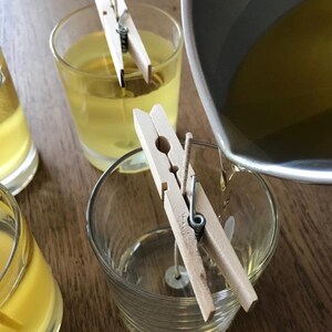 Puede incluir: Primer plano de la fabricaci&oacute;n de velas. Vasos de vidrio transparente llenos de l&iacute;quido amarillo, sujetos con pinzas de madera. Una olla vierte m&aacute;s l&iacute;quido amarillo en un vaso. La escena se desarrolla sobre una superficie de madera.